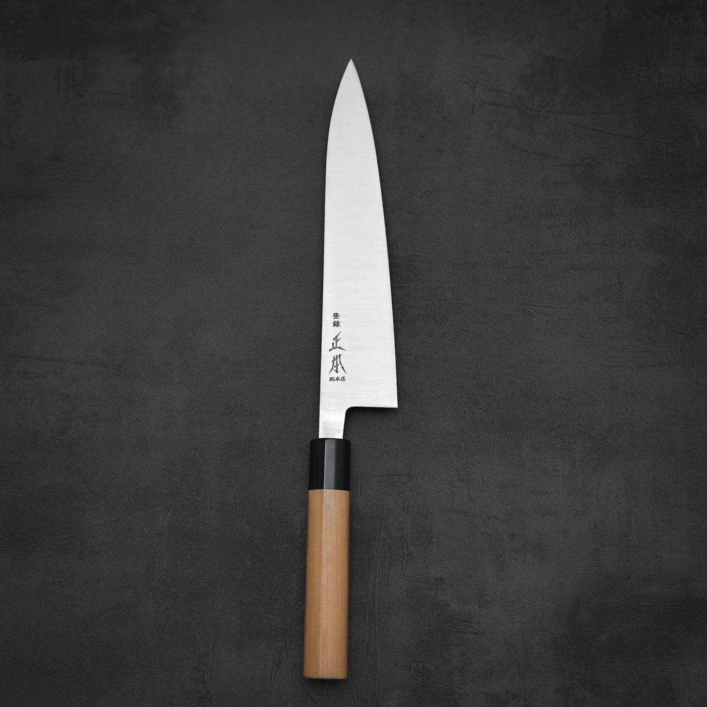 A Masamoto KS gyuto 210mm chef’s knife with a shiny Shirogami#2 steel blade and light wooden handle lies on a dark surface, featuring Japanese characters engraved near the handle.