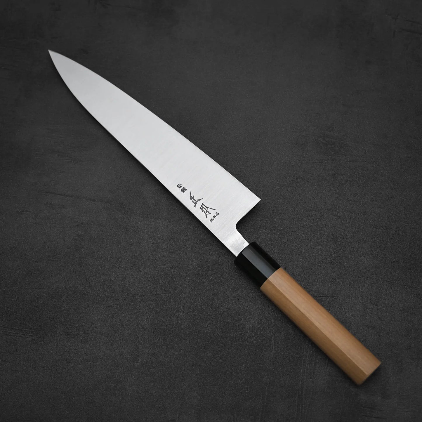 The Masamoto KS gyuto 210mm by Masamoto features a Shirogami#2 steel blade engraved with kanji and a light wood handle in an ambidextrous design, shown on a dark, textured surface.