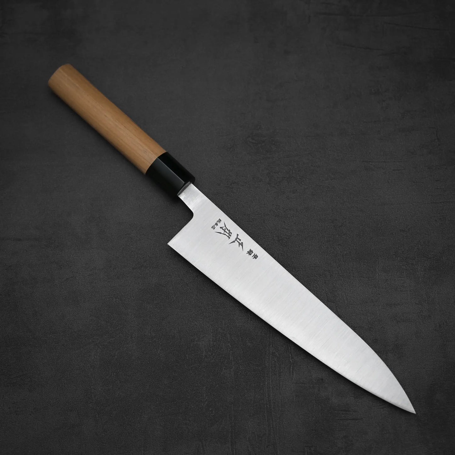 The Masamoto KS gyuto 210mm, a Japanese chefs knife by Masamoto, features a sharp Shirogami#2 steel blade and an ambidextrous wooden handle, shown on a dark textured surface.
