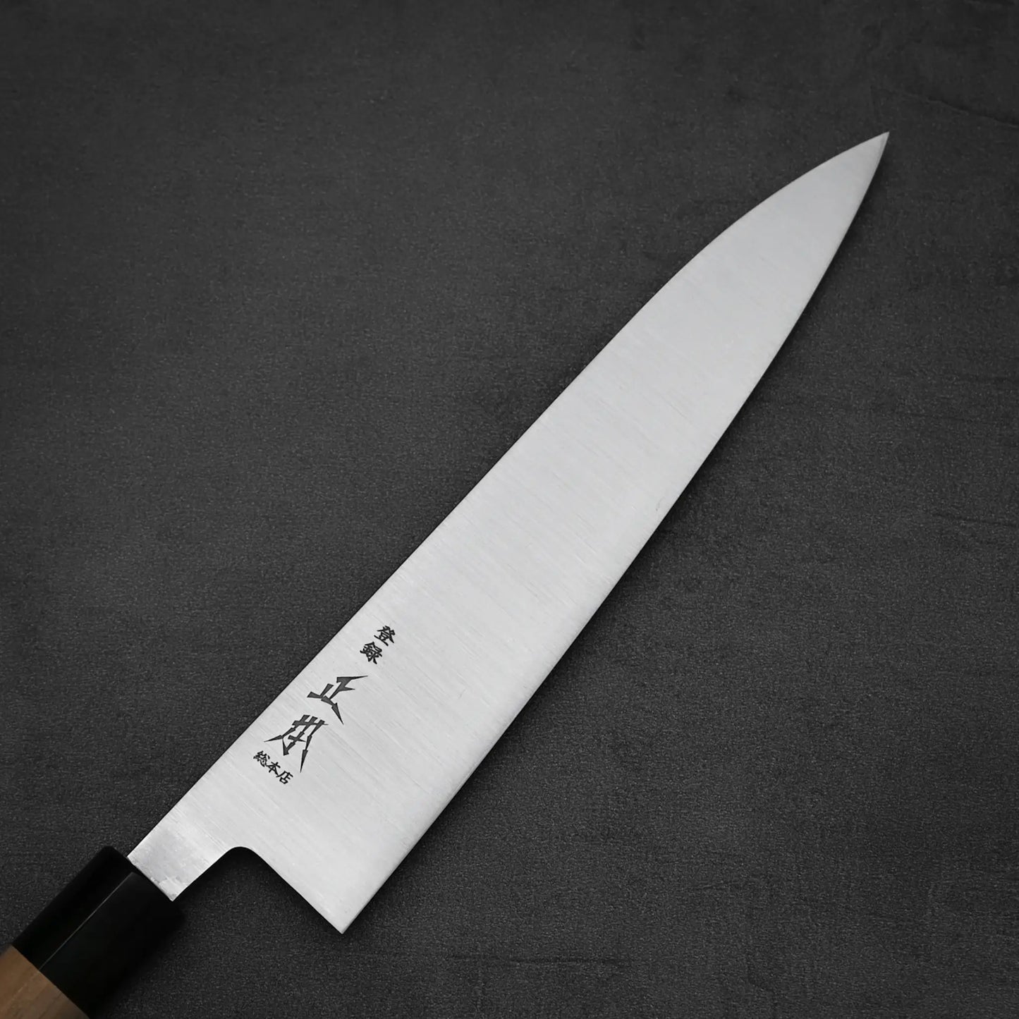 The Masamoto KS gyuto 210mm by Masamoto features a wooden handle and a long, sharp Shirogami#2 steel blade with Japanese characters, displayed on a dark gray textured surface.