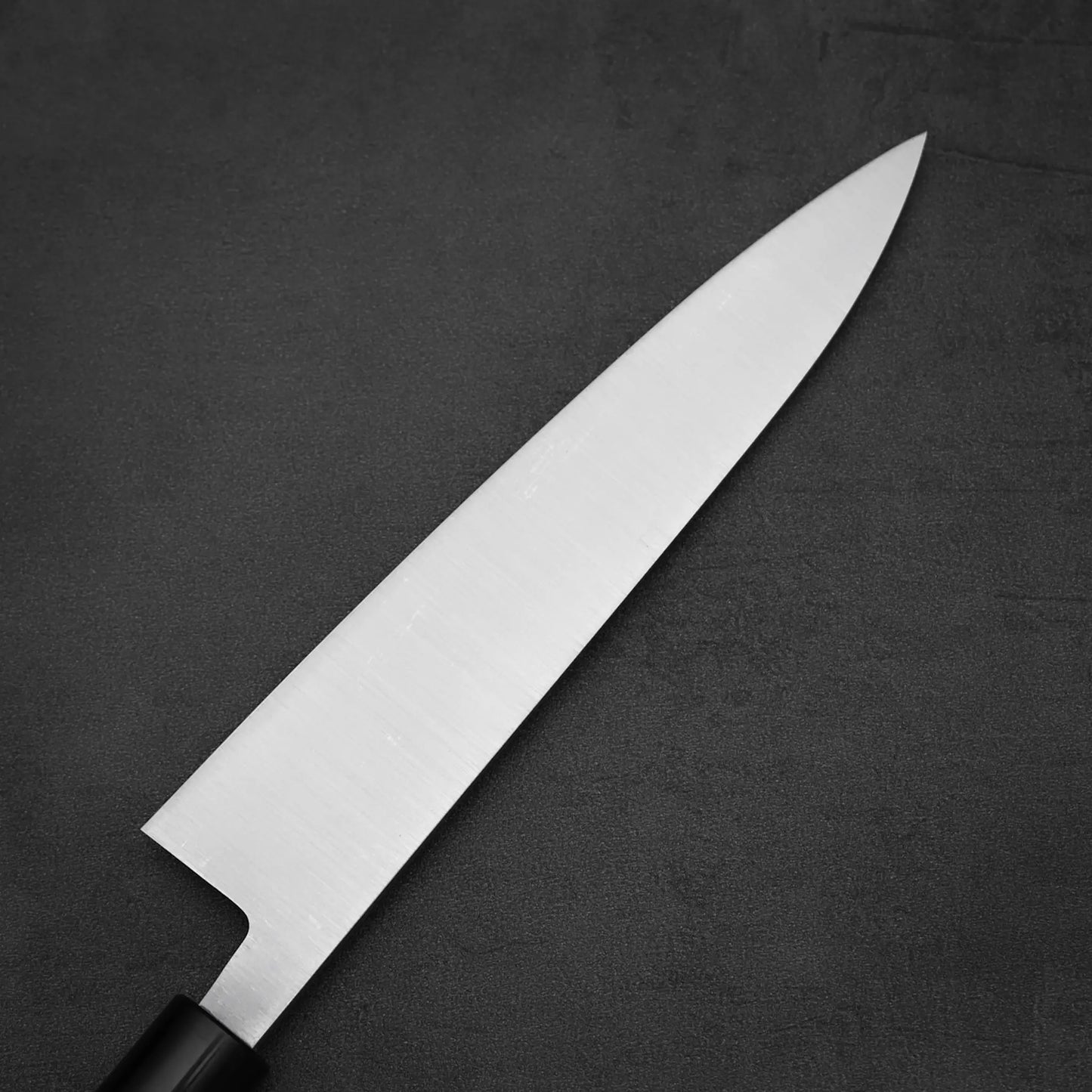 The Masamoto KS gyuto 210mm chefs knife by Masamoto, featuring a clean, pointed Shirogami#2 steel blade and a black handle, lies diagonally from bottom left to top right on a dark textured surface.