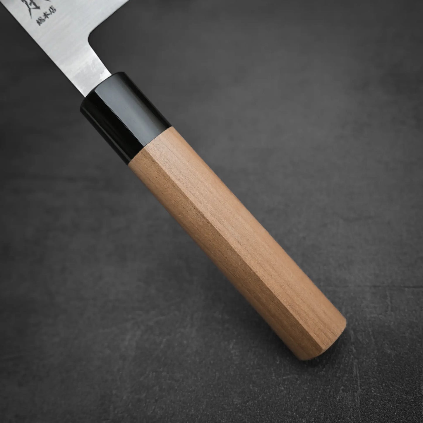 Close-up of a Masamoto KS gyuto 210mm Japanese chefs knife with a wooden handle and Shirogami#2 steel blade, partially shown on a dark, textured surface.