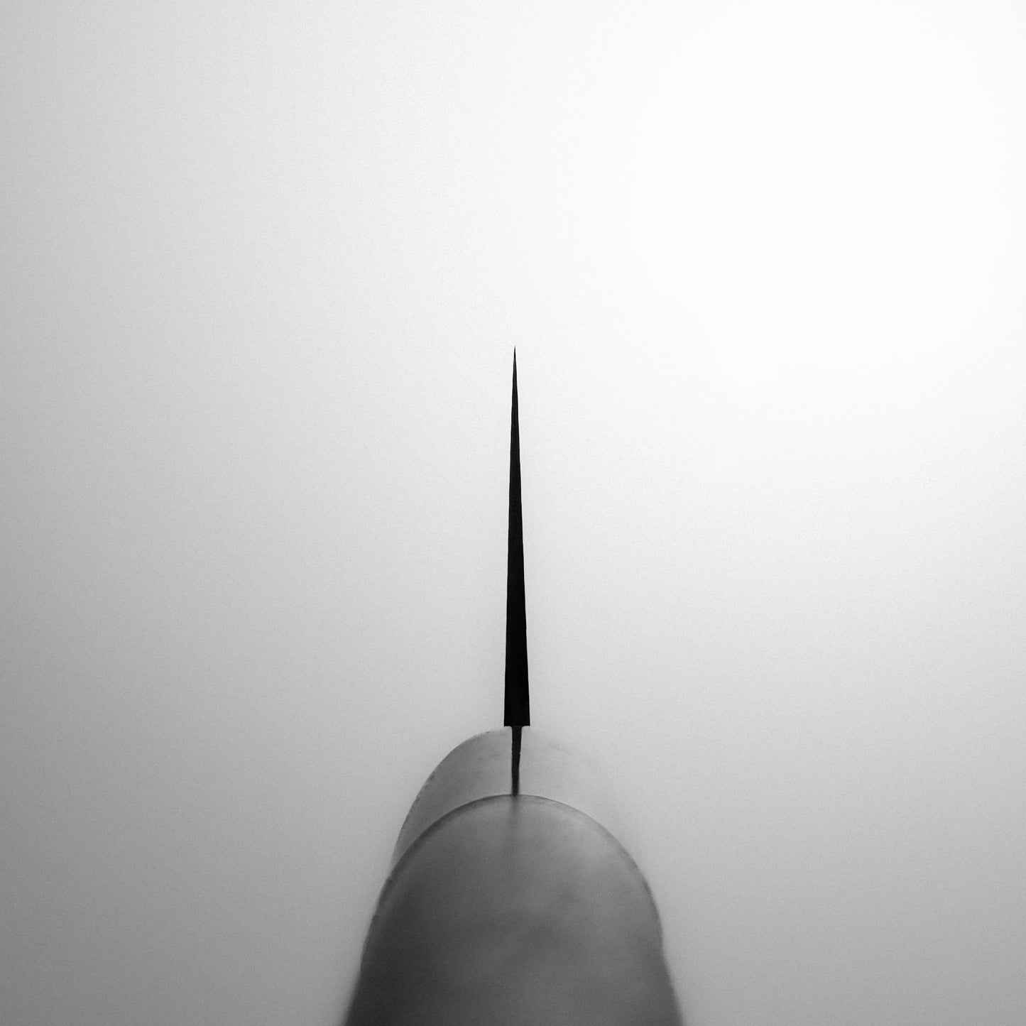 A black metal spire rises from the lower center, evoking a Masamoto KS gyuto 210mm chefs knife. The minimalistic image, in monochrome, features a pale white, foggy background that highlights the sharp elegance of this Masamoto blade.