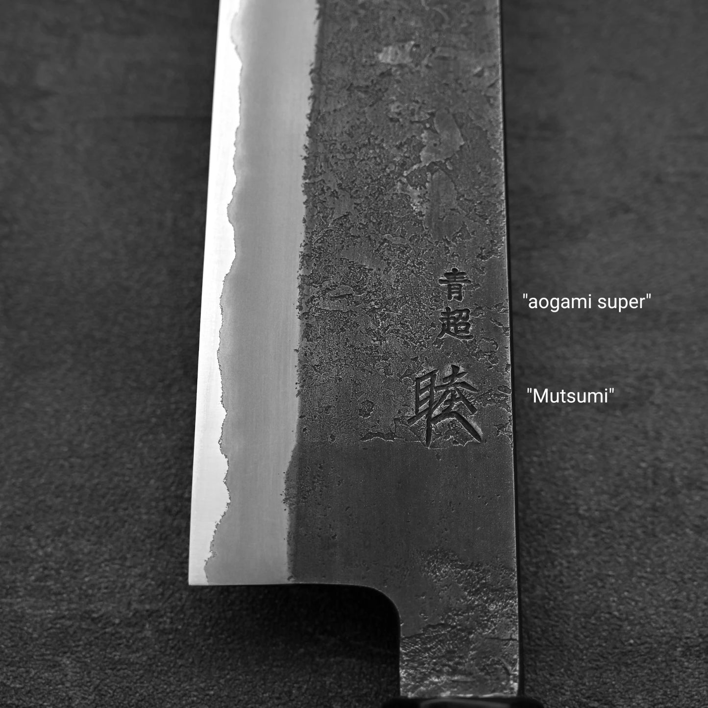 Close-up of the Hinoura kurouchi nashiji AS (stainless clad) gyuto 240mm blade, showing its polished edge, textured upper area, and Japanese kanji characters with “Aogami Super” steel and “Hinoura” visible beside the blade.