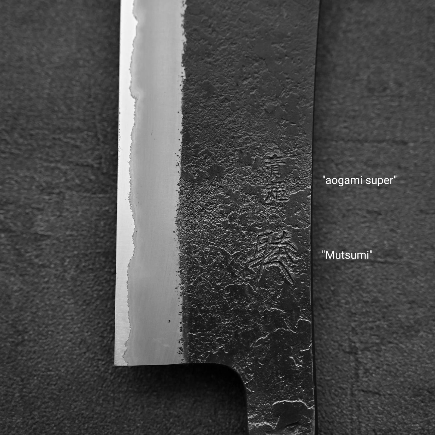 Close-up of the Hinoura kurouchi nashiji AS (stainless clad) hakata 180mm knife blade, showcasing a textured black finish and two Japanese engravings—Aogami Super near the top, Mutsumi below—against a dark background.