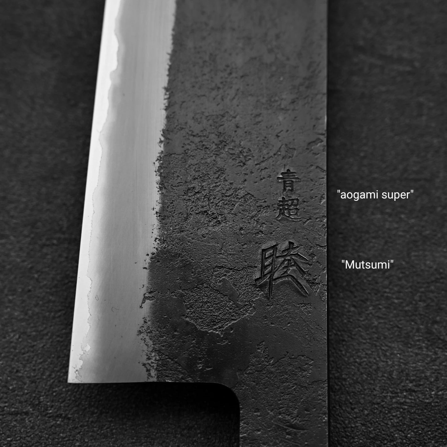 Close-up of the Hinoura kurouchi nashiji AS (stainless clad) kiritsuke gyuto 210mm blade, featuring a textured dark finish and engraved characters labeled with English translations Aogami Super steel and Mutsumi.