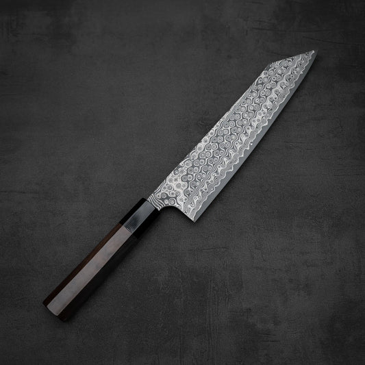 The Nigara Hamono Nigara Anmon SG2 Damascus kiritsuke gyuto 240mm, featuring a stunning Damascus steel blade and dark wood handle, is showcased on a dark textured surface.