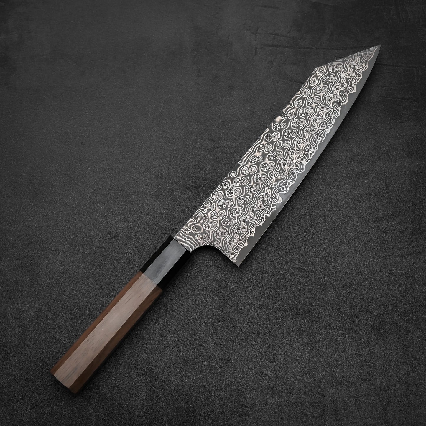 The Nigara Hamono Nigara Anmon SG2 damascus kiritsuke gyuto 210mm, featuring a striking damascus blade and wooden handle, rests on a dark textured surface.
