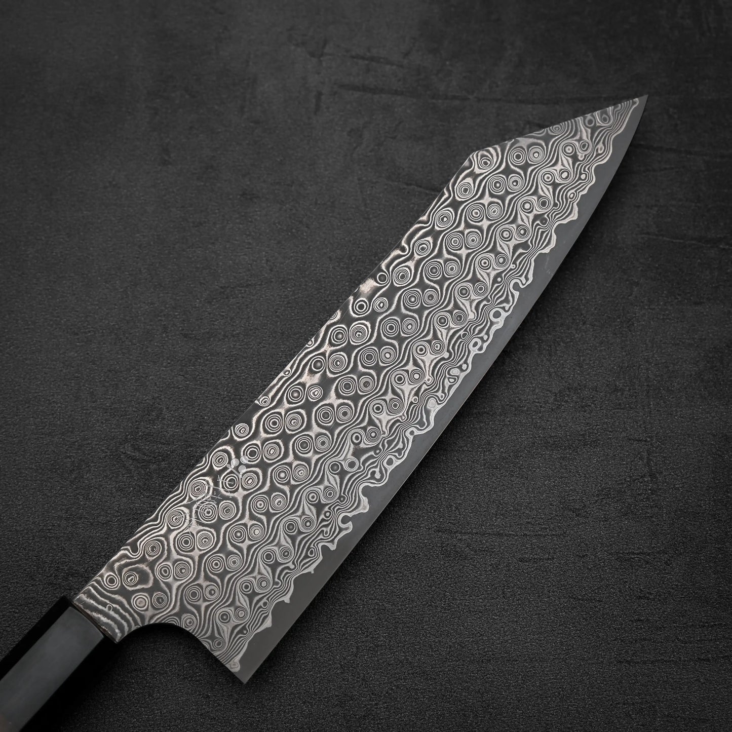 A close-up of the Nigara Hamono Nigara Anmon SG2 damascus kiritsuke gyuto 210mm, showcasing its wide, pointed blade with an intricate circular damascus pattern, crafted from premium SG2 steel on a dark textured surface.