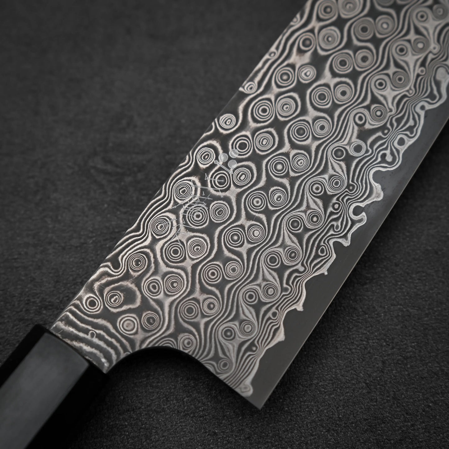 A close-up of the Nigara Hamono Nigara Anmon SG2 damascus kiritsuke gyuto 210mm knife with a black handle, showcasing its intricate damascus patterns on the blade, set against a dark background.