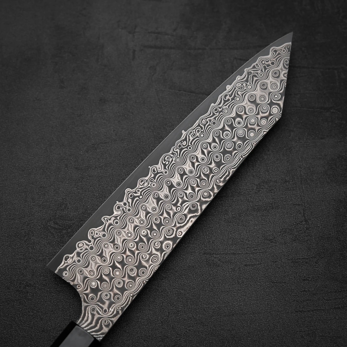 The Nigara Hamono Nigara Anmon SG2 damascus kiritsuke gyuto 210mm features a stunning swirling damascus pattern blade, beautifully displayed on a dark, textured surface.