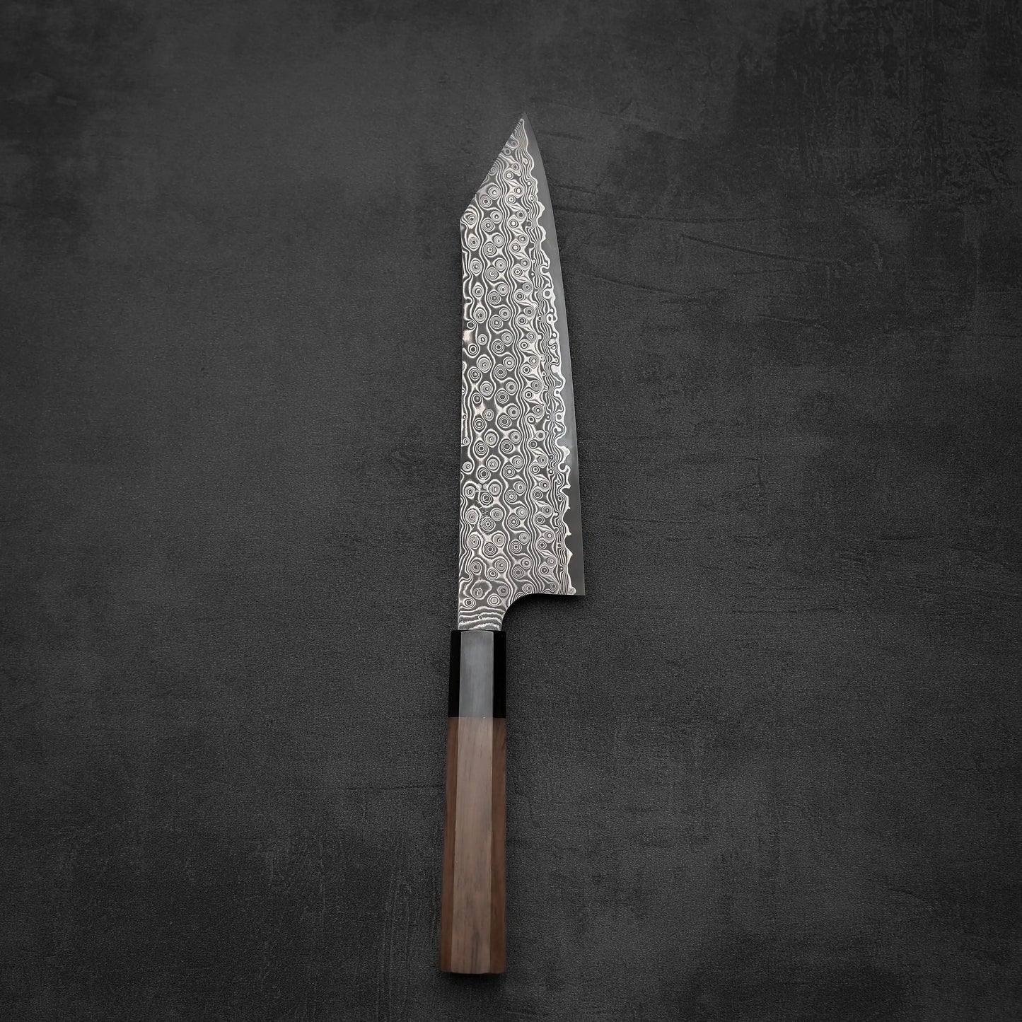 The Nigara Hamono Nigara Anmon SG2 damascus kiritsuke gyuto 210mm, featuring a wooden handle and striking damascus pattern, rests on a dark textured surface.
