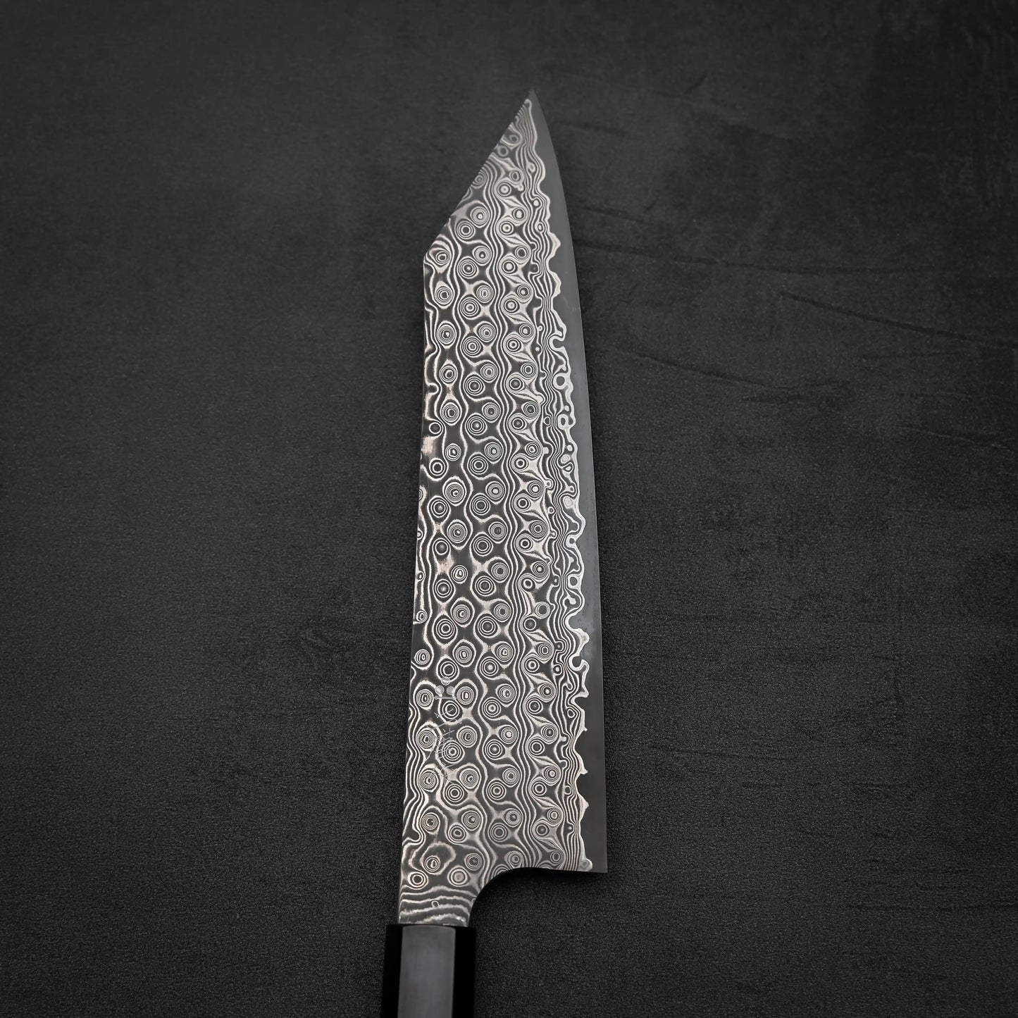 A close-up of the Nigara Hamono Nigara Anmon SG2 Damascus kiritsuke gyuto 210mm highlights its stunning Damascus blade and black handle, set against a dark textured background.