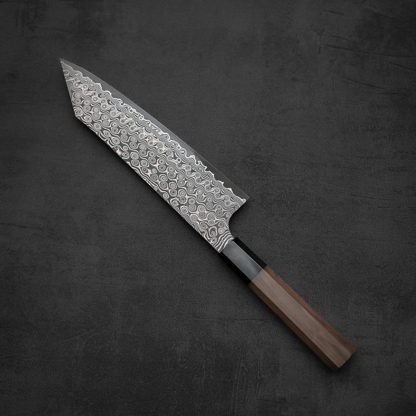 The Nigara Hamono Nigara Anmon SG2 damascus kiritsuke gyuto 210mm, featuring a striking Damascus-patterned SG2 steel blade and a wooden handle, rests on a dark textured surface.
