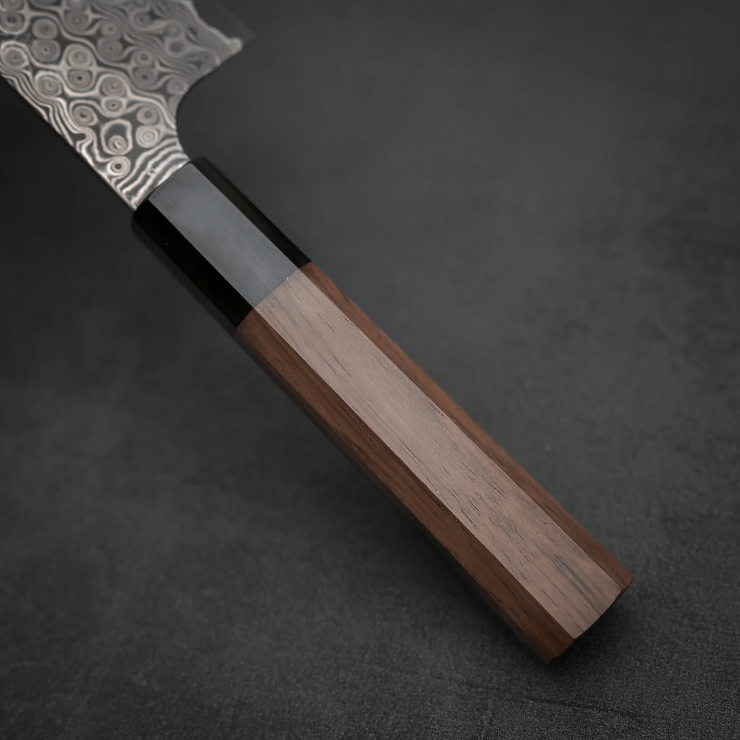 Close-up of the Nigara Hamono Nigara Anmon SG2 damascus kiritsuke gyuto 210mm, showing its polished wood handle, black bolster, and part of the SG2 steel damascus blade against a dark textured background.