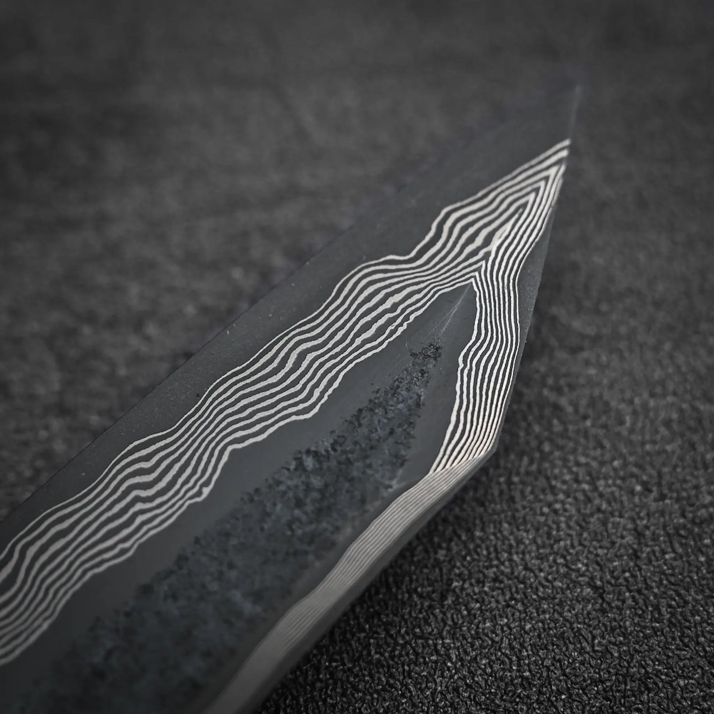Close-up of the Nigara Hamono Nigara kurozome ginsan damascus kiritsuke petty knife 150mm blade tip, showing its wavy Damascus steel pattern against a textured dark background.