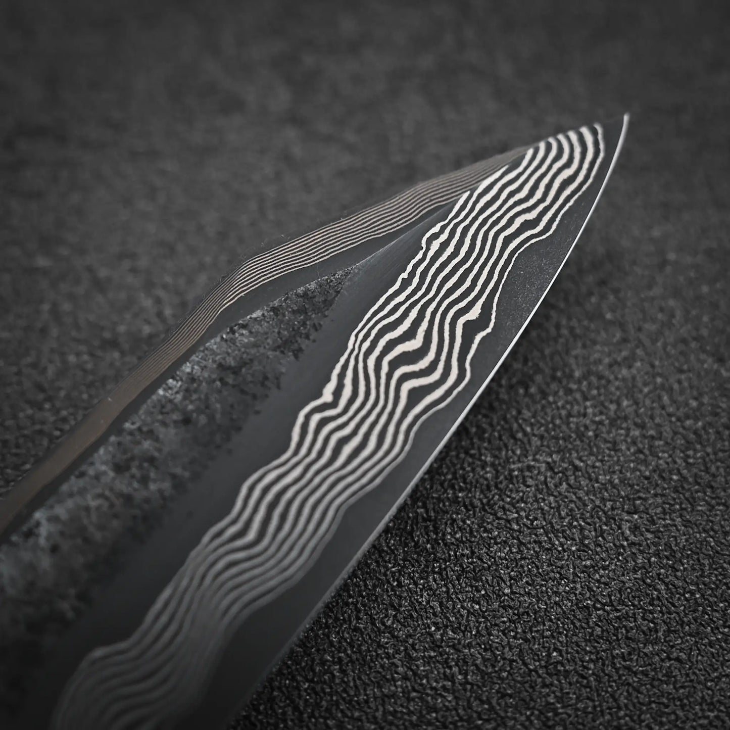 Close-up of the Nigara Hamono Nigara kurozome ginsan damascus kiritsuke petty knife 150mm blade, showcasing its unique wavy Damascus pattern against a textured black background.