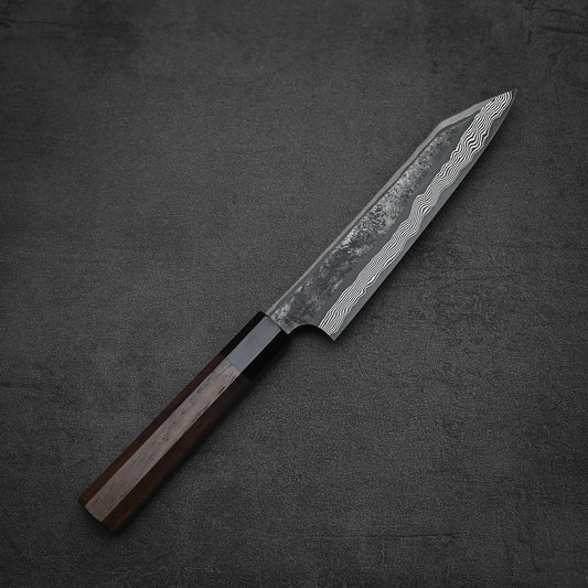 The Nigara Hamono Nigara kurozome ginsan damascus kiritsuke petty knife 150mm, featuring a striking Damascus blade and dark wooden handle, is displayed on a textured dark gray surface.