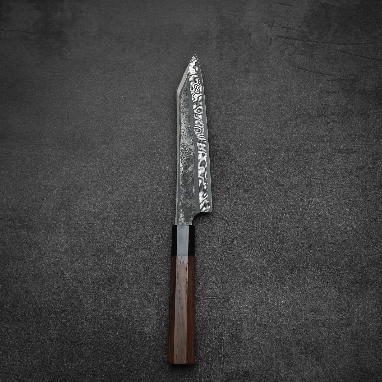 The Nigara Hamono Nigara kurozome ginsan damascus kiritsuke petty knife 150mm, featuring a Damascus pattern blade and dark wooden handle, is displayed on a dark, textured surface.