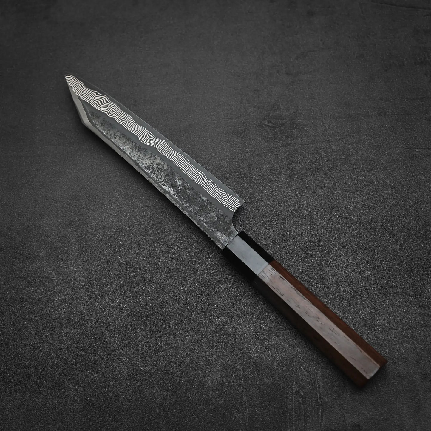 The Nigara Hamono Nigara kurozome ginsan damascus kiritsuke petty knife 150mm features a straight-edged, Damascus-patterned blade with wavy lines near the spine and a dark handle, resting on a dark textured surface.