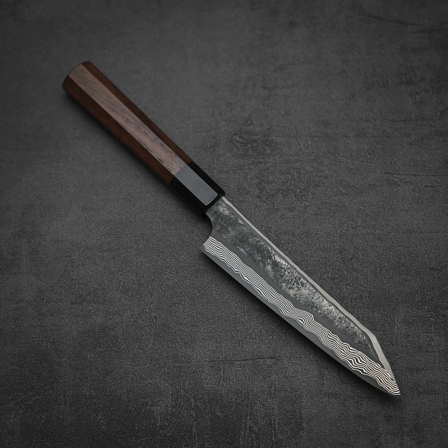 The Nigara Hamono Nigara kurozome ginsan damascus kiritsuke petty knife 150mm, featuring a dark wooden handle and a striking Damascus steel blade, rests on a dark gray textured surface.