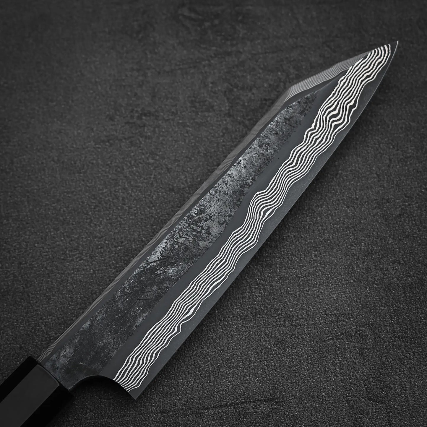 A close-up of the Nigara Hamono Nigara kurozome ginsan damascus kiritsuke petty knife 150mm blade, featuring a distinct Damascus pattern along the edge against a dark, textured background.