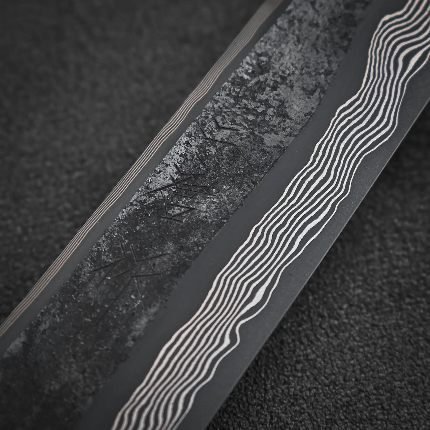 Close-up of the Nigara kurozome ginsan damascus kiritsuke petty knife 150mm by Nigara Hamono, showcasing its textured, dark matte blade with wavy Damascus pattern, set against a black grainy background.