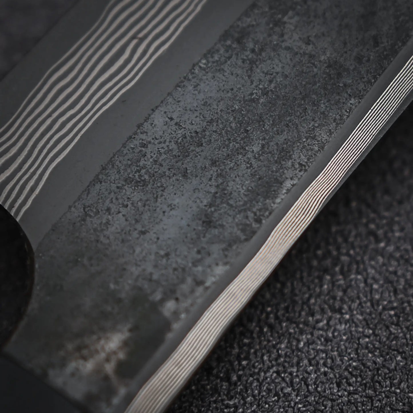 Close-up of the Nigara Hamono Nigara kurozome ginsan damascus kiritsuke petty knife 150mm blade, with its textured, worn Ginsan steel surface and fine wavy Damascus lines, shown against a dark, rough background.