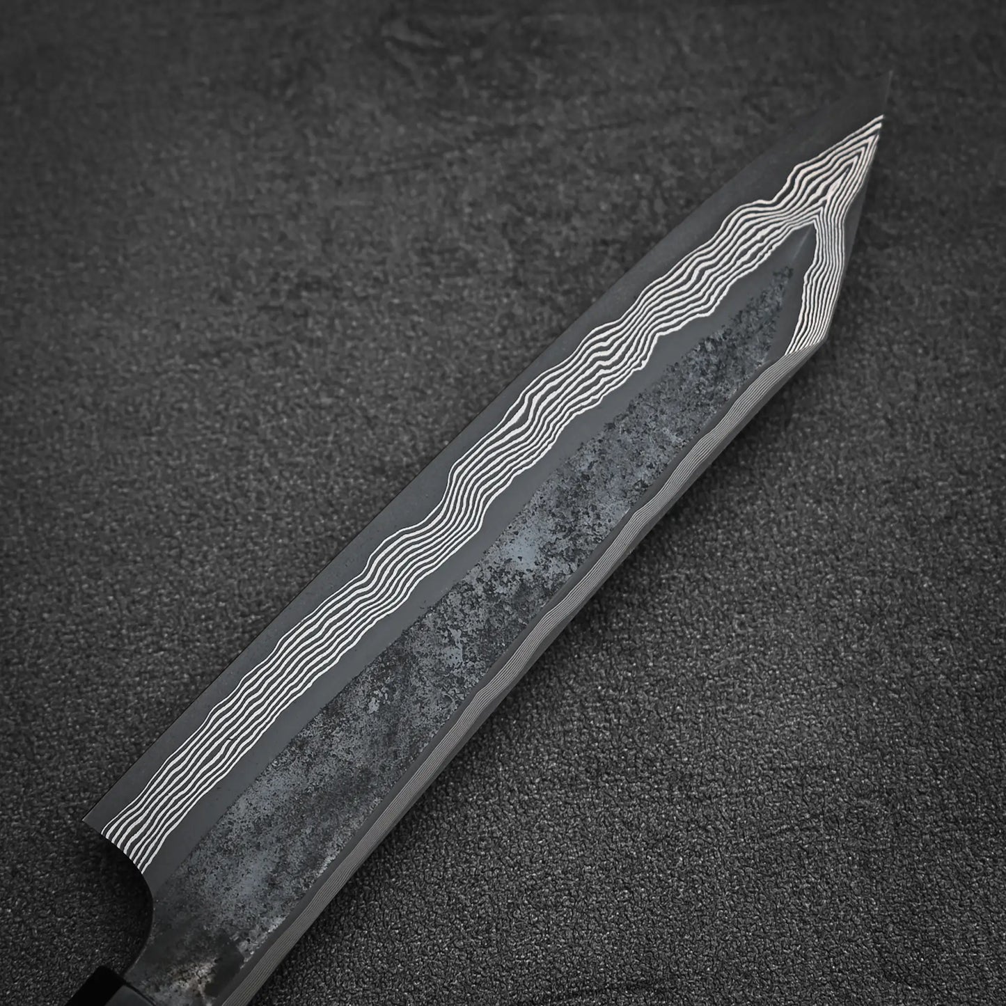 A close-up of the Nigara Hamono Nigara kurozome ginsan damascus kiritsuke petty knife 150mm blade, highlighting its wavy Damascus patterns and textured black finish against a dark background.