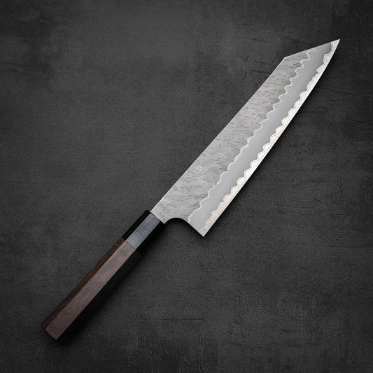 The Nigara Hamono Nigara tsuchime AS kiritsuke gyuto 240mm, featuring a patterned steel blade and dark wooden handle, is elegantly displayed on a dark textured surface.