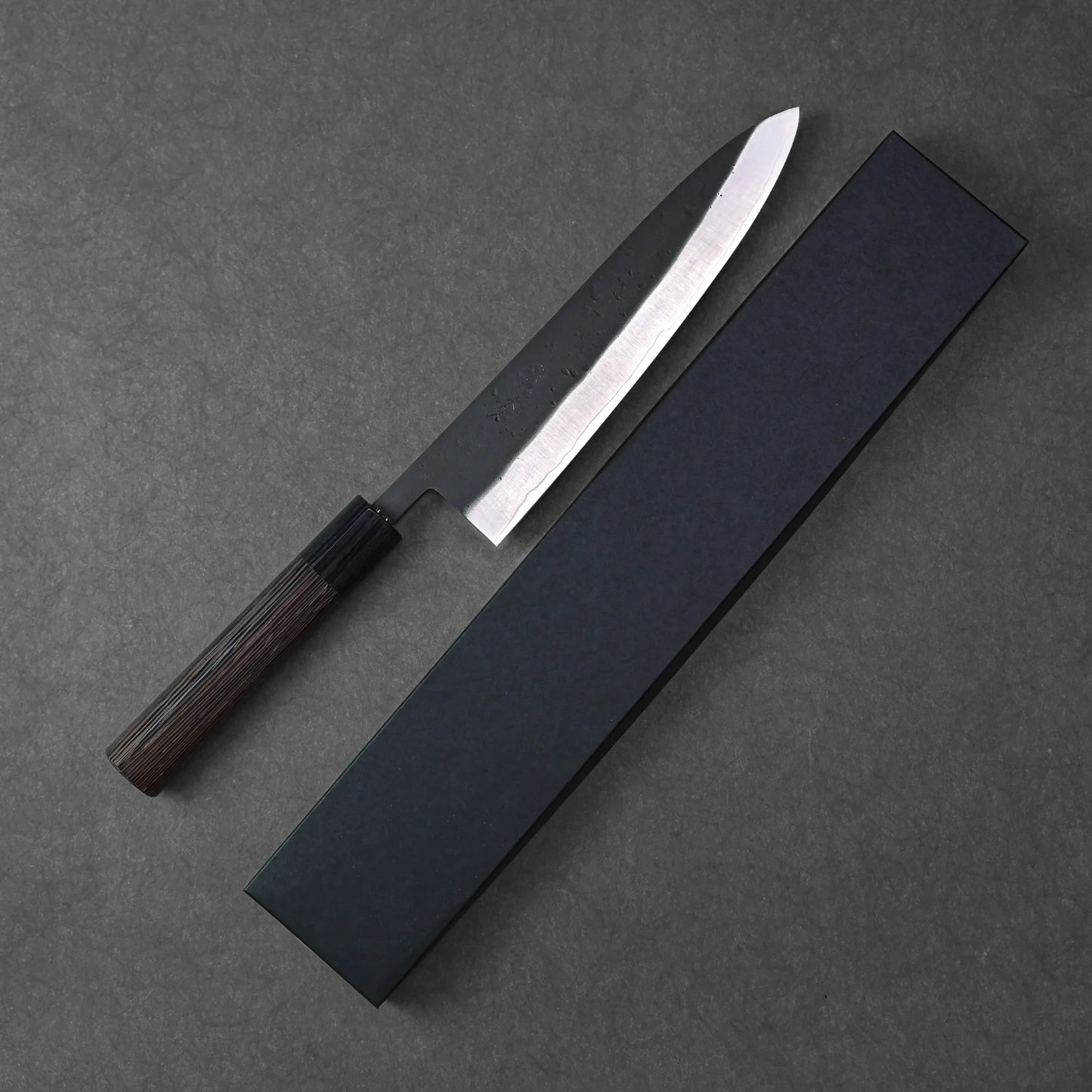 The Ohishi kurouchi SLD gyuto 210mm, featuring a dark wooden handle and black blade, rests beside its rectangular black box on a dark textured surface.