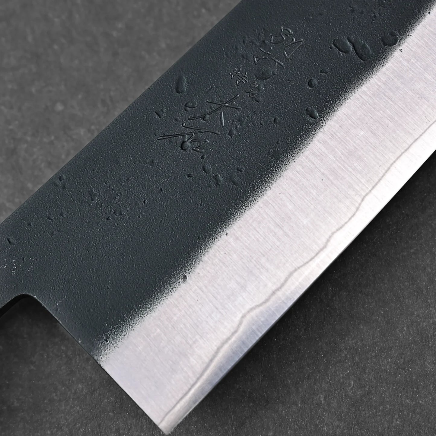 Close-up of the Ohishi kurouchi SLD gyuto 210mm knife blade by Ohishi, featuring a polished edge, textured upper section with engraved Japanese characters and water droplets, showcasing its craftsmanship.