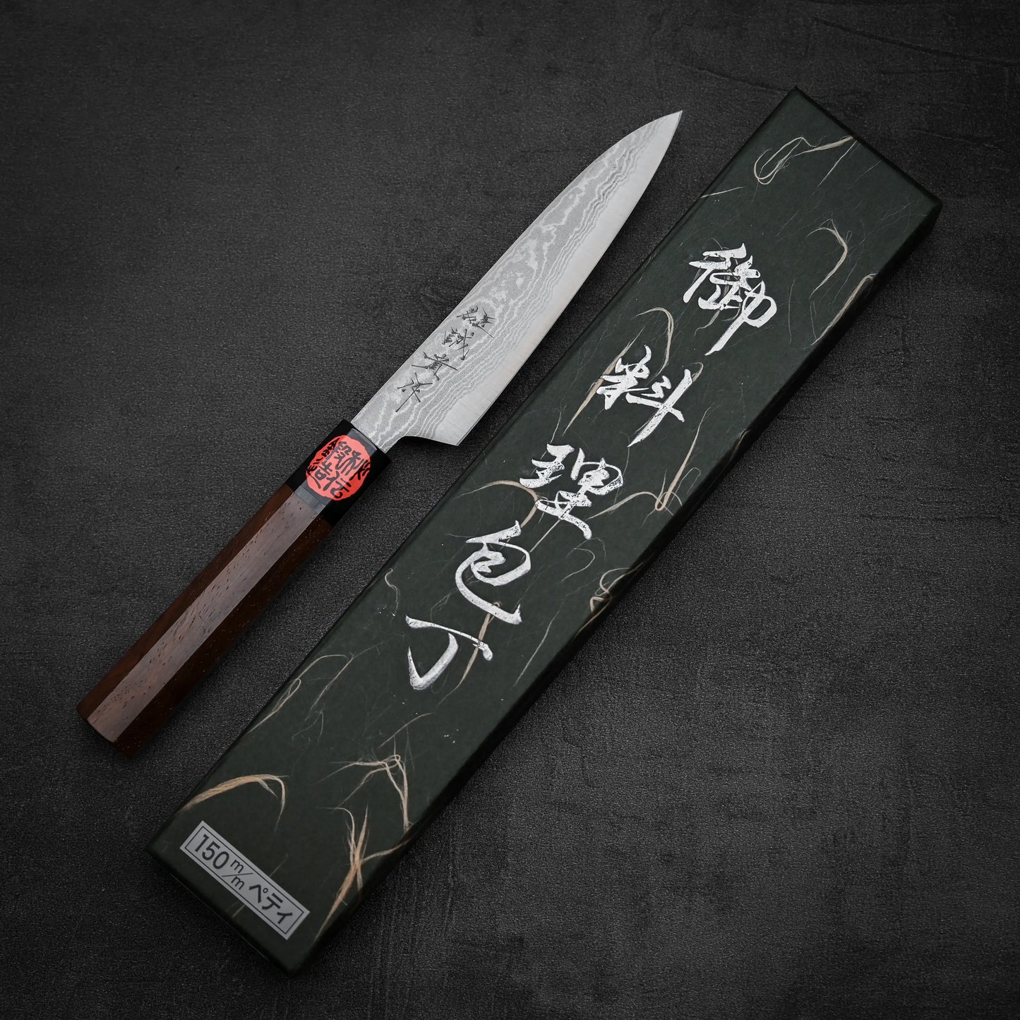 A Shigeki Tanaka aogami#2 damascus petty knife with a patterned blade and wooden handle is displayed next to a black box featuring white Japanese text on the lid, set against a dark textured background.