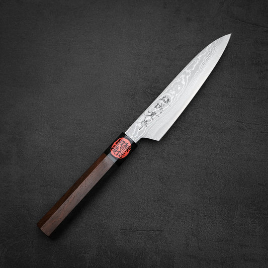 A Shigeki Tanaka aogami#2 damascus petty knife with a wooden handle, engraved patterns, and a red and black label near the handle rests on a dark textured surface.