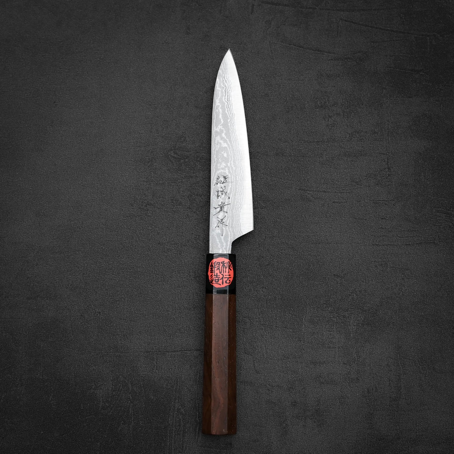 The Shigeki Tanaka aogami#2 damascus petty knife features a patterned blade with engraved markings, a wooden handle with a red accent, and rests on a dark textured surface.