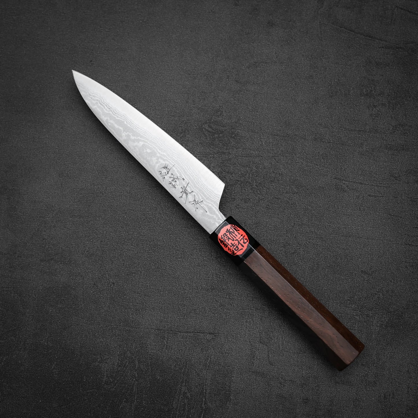 The Shigeki Tanaka aogami#2 damascus petty knife features a patterned blade with engraved characters and a dark wooden handle with a red label, displayed on a dark textured surface.