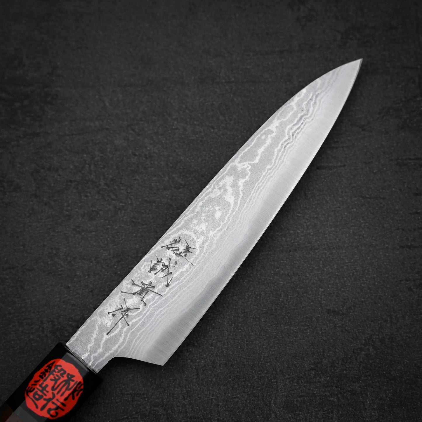 A Shigeki Tanaka aogami#2 damascus petty knife with a patterned blade, kanji engraving, and a black handle with red cap rests on a dark textured surface.