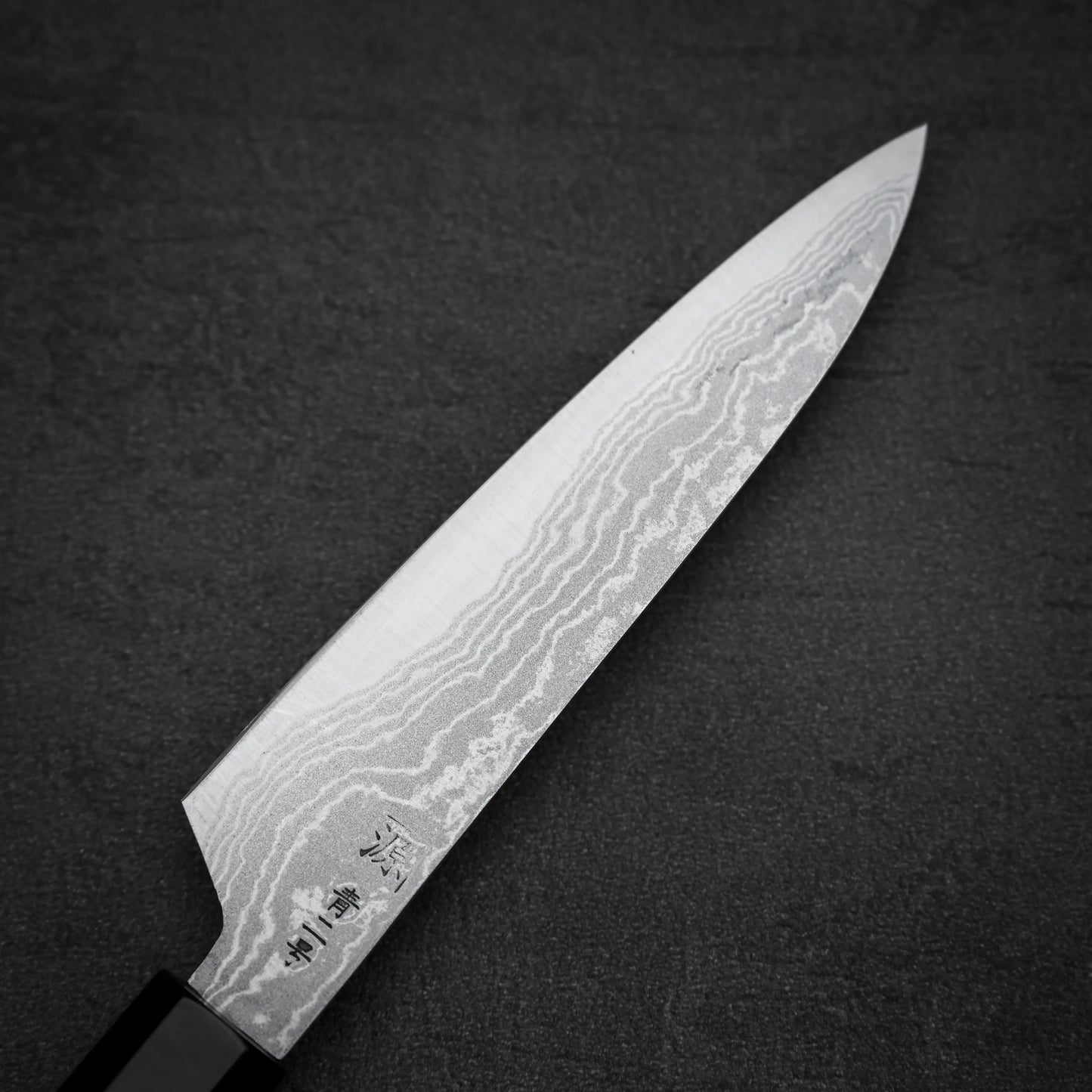The Shigeki Tanaka aogami#2 damascus petty knife, featuring a patterned blade and black handle, is shown on a dark textured surface. Japanese characters are engraved near the blades base.
