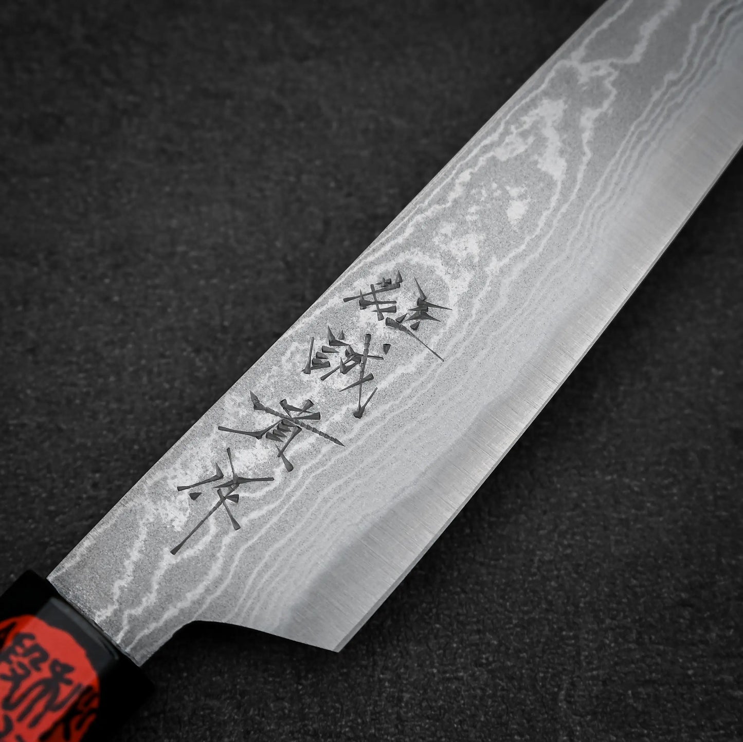 A close-up of the Shigeki Tanaka aogami#2 damascus petty knife shows distinct Damascus steel patterns and Japanese characters engraved on the blade, set against a dark textured surface.