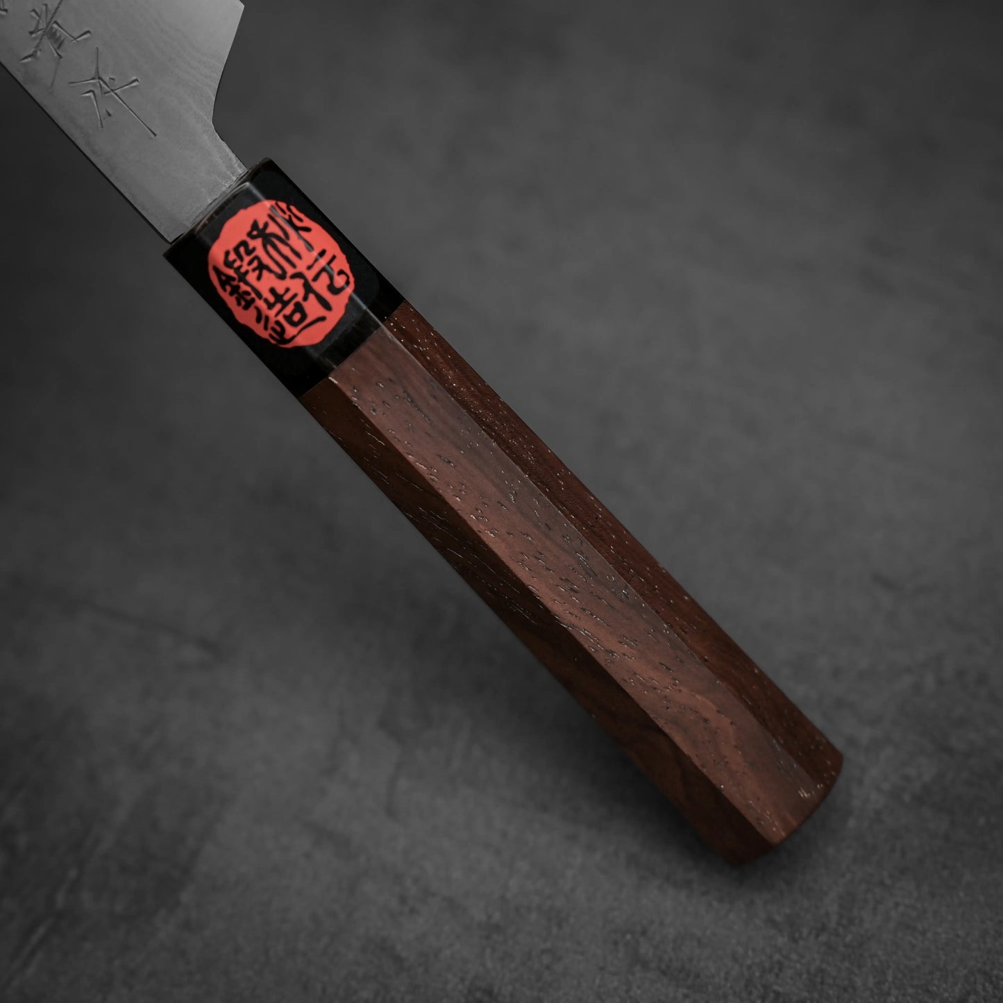 Close-up of the Shigeki Tanaka aogami#2 damascus petty knife handle, showing its hexagonal shape, black ferrule, and a red seal with black Japanese characters near the blade on a smooth dark grey background.