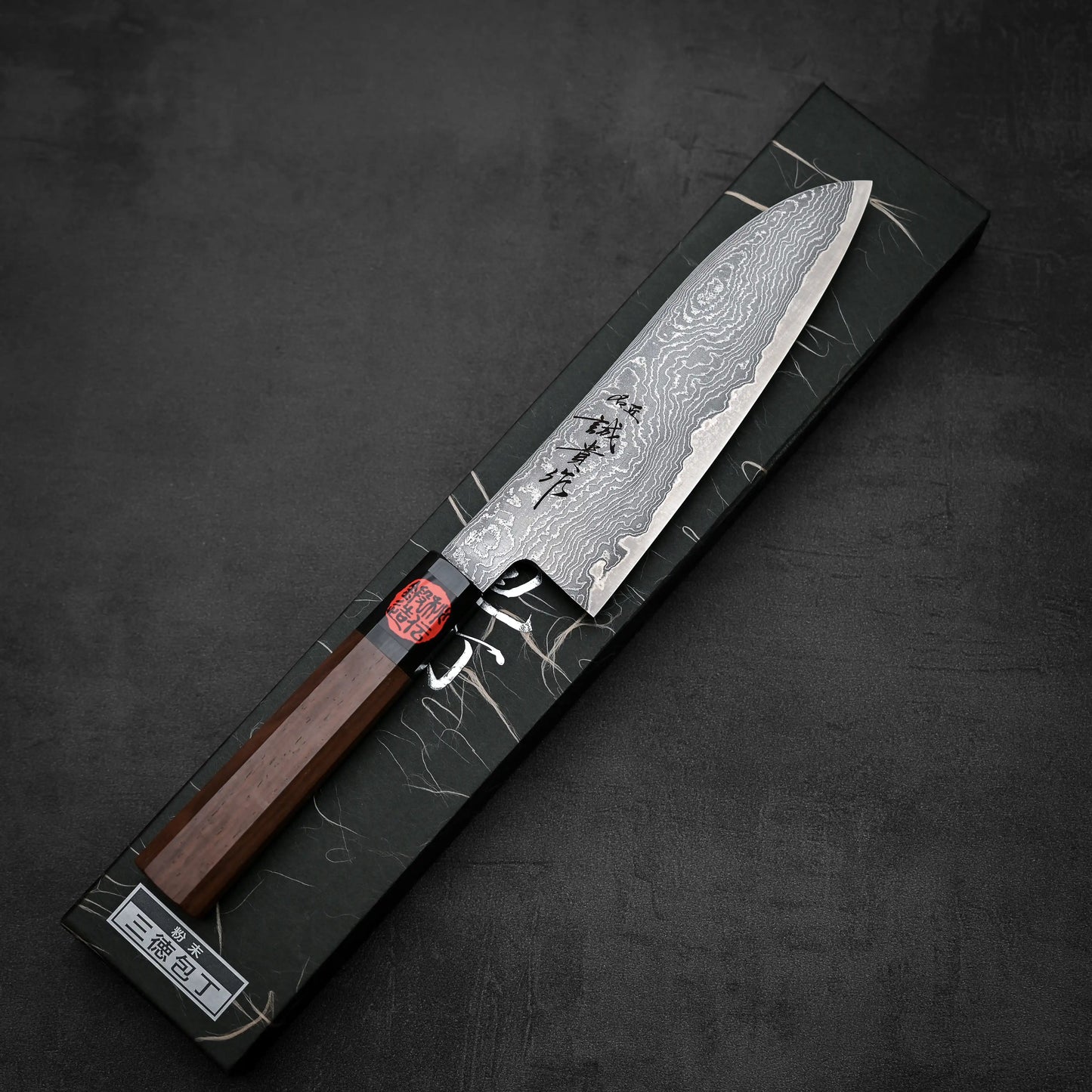 The Shigeki Tanaka R2 damascus santoku 170mm, featuring a wooden handle, is shown atop a decorative black box with subtle patterns and Japanese characters.