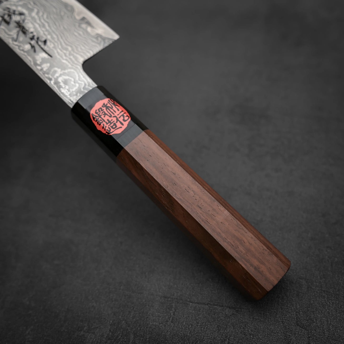 Close-up of the Shigeki Tanaka R2 damascus santoku 170mm knife with a patterned blade and dark wooden handle, resting on a textured gray surface. A red and black label with Japanese characters is near the blade.