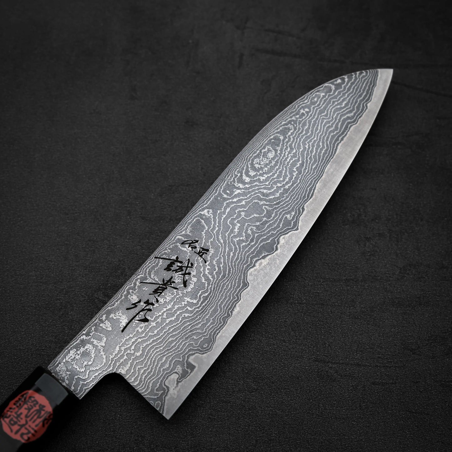 A close-up of the Shigeki Tanaka R2 damascus santoku 170mm knife, featuring a patterned blade with engraved Japanese characters and a dark handle, displayed on a black textured surface.