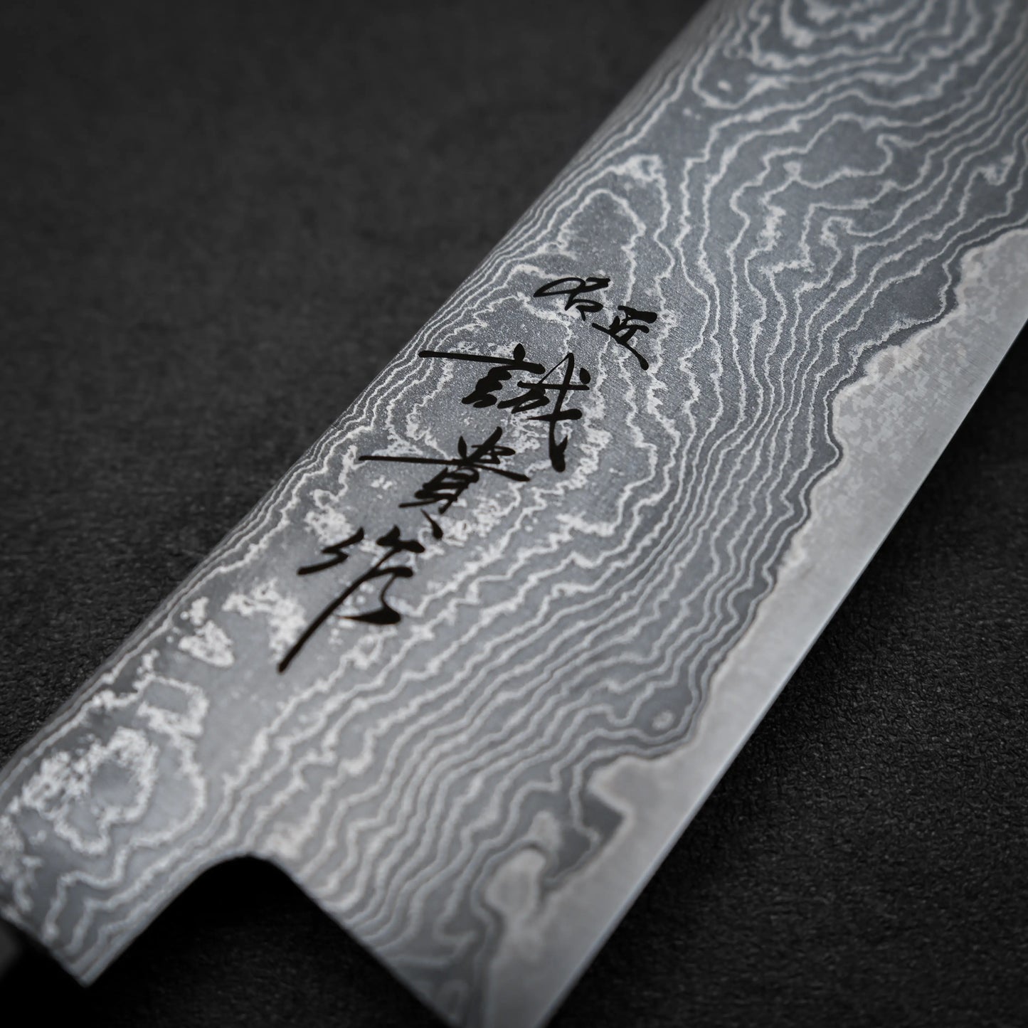 Close-up of the Shigeki Tanaka R2 damascus santoku 170mm blade featuring striking damascus steel patterns and black Japanese characters etched on its surface, set against a dark background.