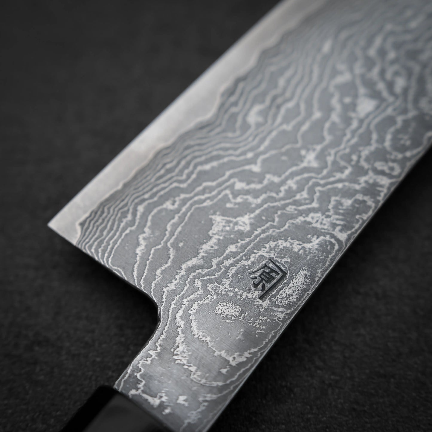 Close-up of the Shigeki Tanaka R2 damascus santoku 170mm blade, featuring striking damascus steel patterns and a small Japanese character near the handle, set against a dark background.
