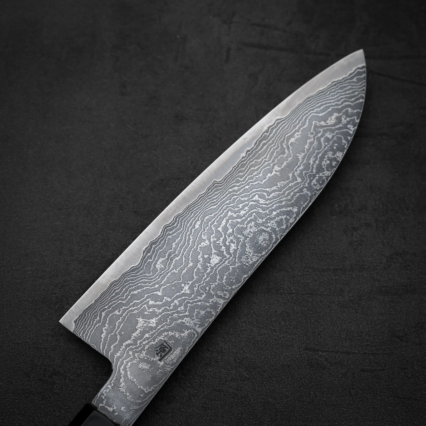 The Shigeki Tanaka R2 damascus santoku 170mm, featuring a beautiful Damascus steel blade, rests on a dark, textured surface.