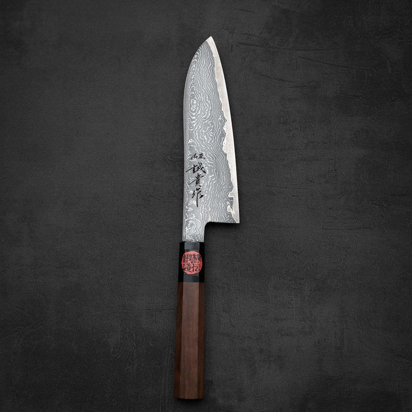 The Shigeki Tanaka R2 damascus santoku 170mm features a striking Damascus steel blade, engraved Japanese characters, and a wooden handle, set against a dark textured background.