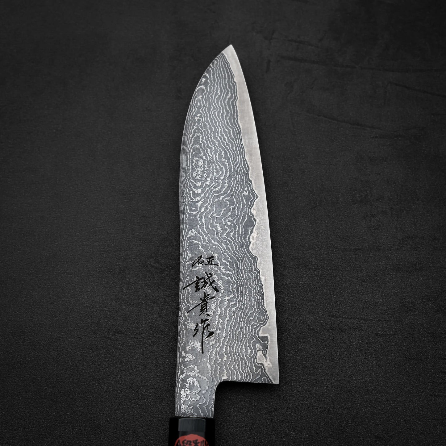 The Shigeki Tanaka R2 Damascus Santoku 170mm features a patterned blade with Japanese characters, set on a dark textured surface.