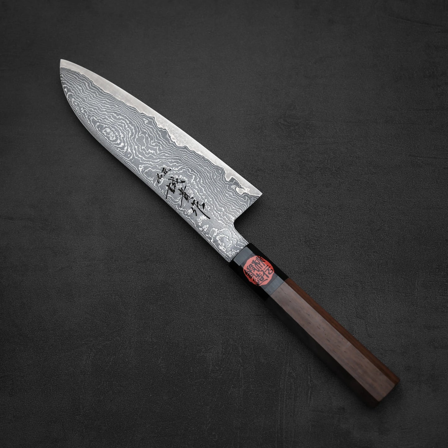 The Shigeki Tanaka R2 damascus santoku 170mm features a wavy-patterned R2 Damascus steel blade with Japanese characters, set on a dark textured surface. The handle is dark wood with a black section and a red seal.