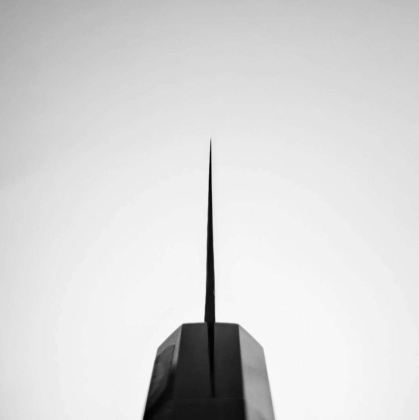 A black and white photo shows the sharp tip of a tall, narrow structure resembling the blade of a Shigeki Tanaka R2 damascus santoku 170mm knife against a clear sky, with its base at the bottom of the image.